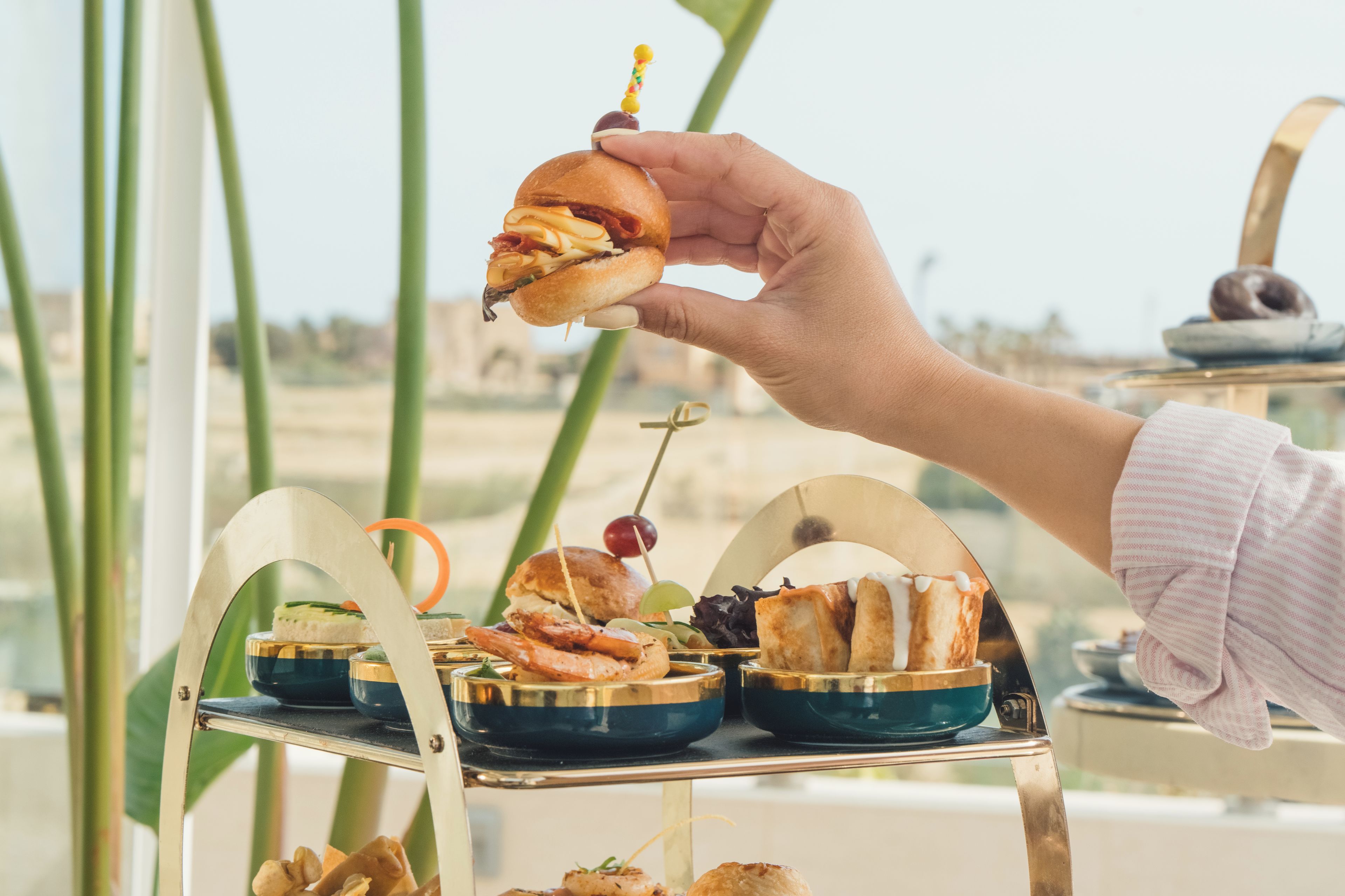 A hand holding a sandwich with a toothpick, with a buffet table in the background.