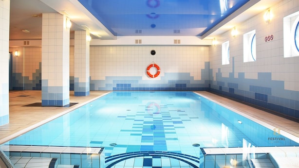 An indoor swimming pool with a blue and white tiled floor, a central lifebuoy, and a ceiling with a blue pattern. - Hotel Festival (Opole)