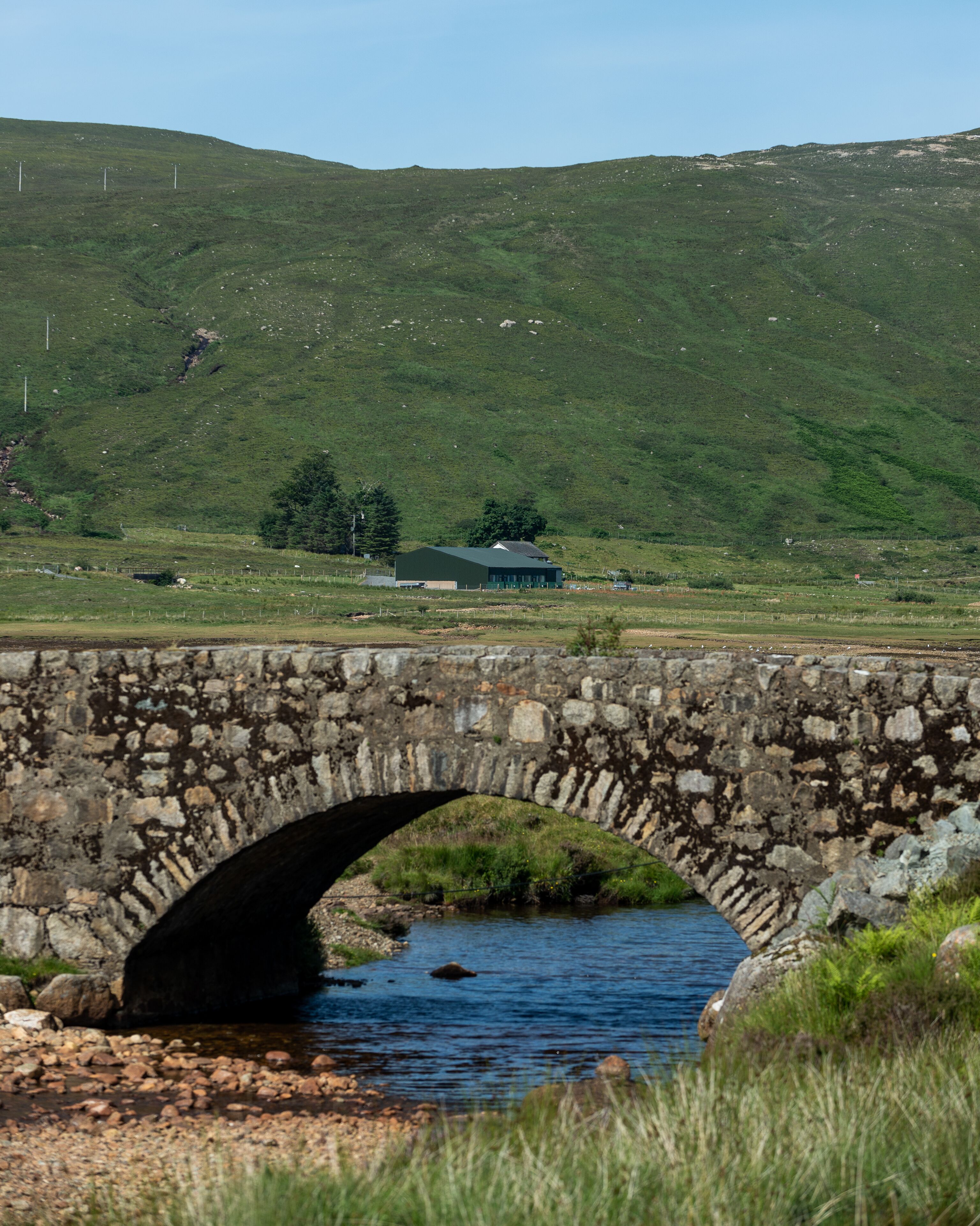 A stone bridge over a river with a green landscape and a building in the background.