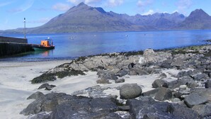 A rocky shoreline with a boat docked at a pier, a mountain in the background, and calm water. - Kinloch Ainort Apartments  (Sconser)