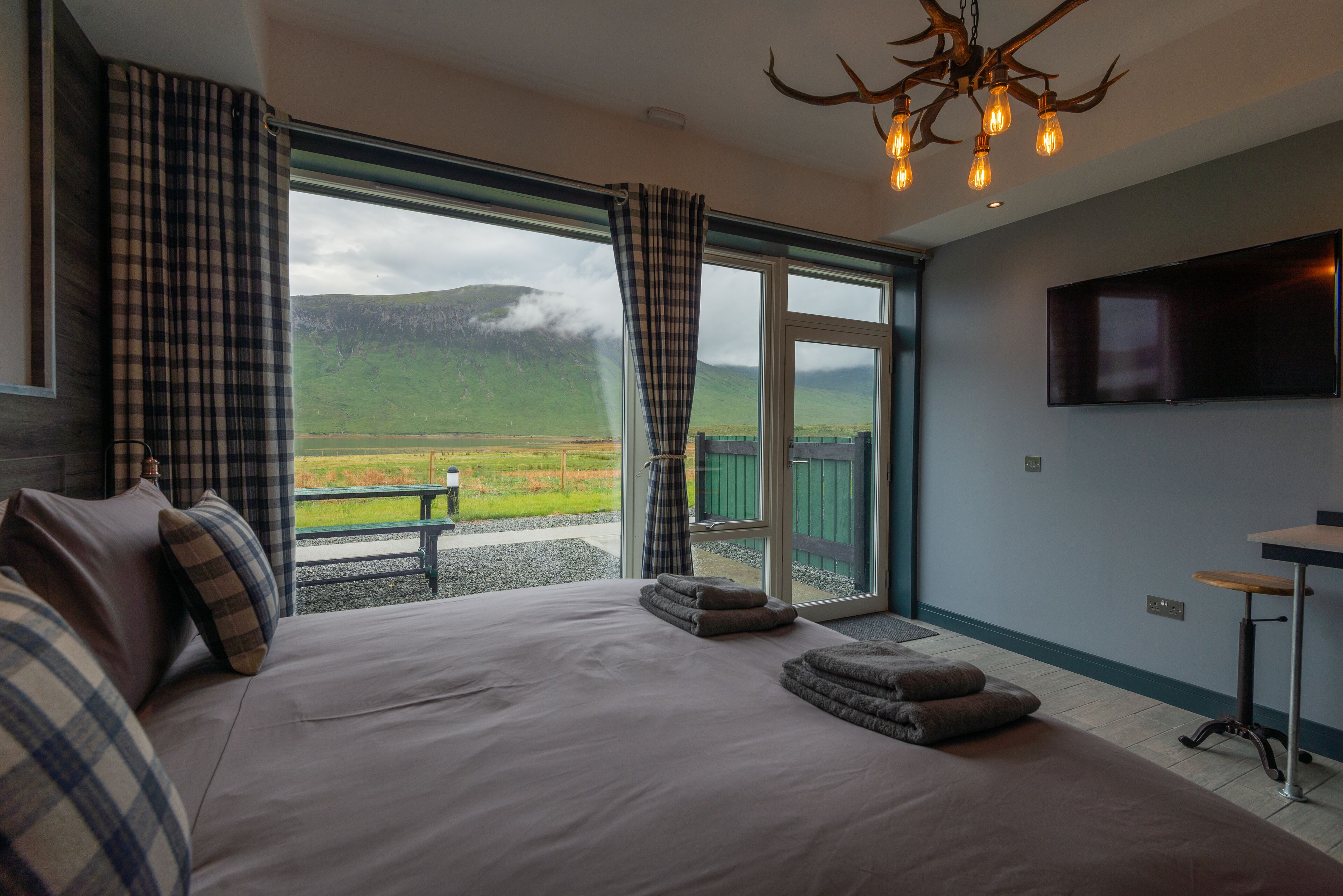 A bedroom with a large bed, plaid curtains, a television, and a view of a green landscape.