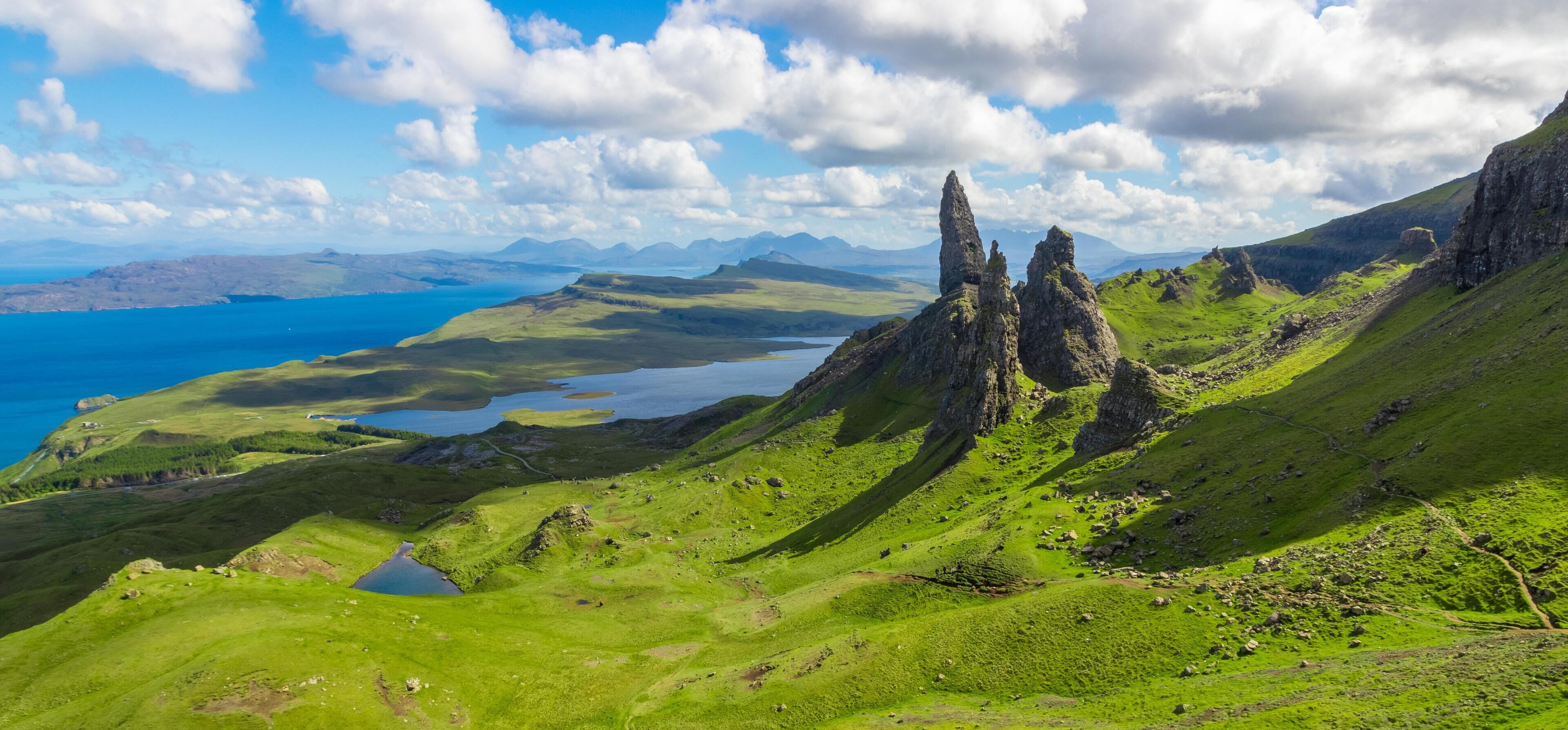 Une vue panoramique d’un paysage montagneux, avec des collines verdoyantes, un lac et des montagnes lointaines, le tout sous un ciel partiellement nuageux.