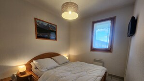 A bedroom with a wooden bed, a nightstand, a lamp, a picture on the wall, a window, and a ceiling light. - Cozy 4-person apartment with balcony & garage in Samoëns (Samoëns)