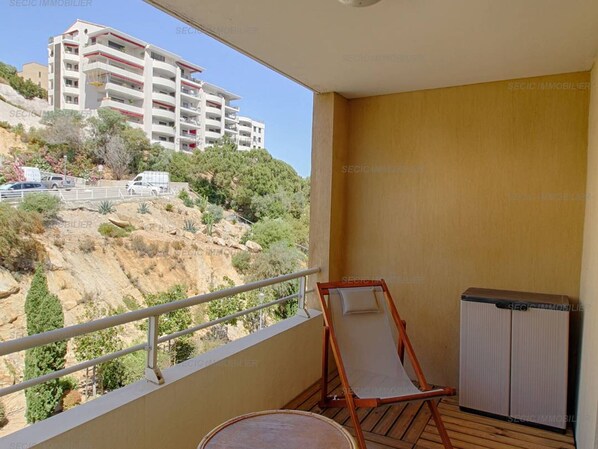 A balcony with a view of a hillside and a building. There is a wooden chair and a small table.