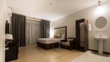 A modern hotel room with a bed, a chair, a desk, a mirror, a sink, and a window with curtains.