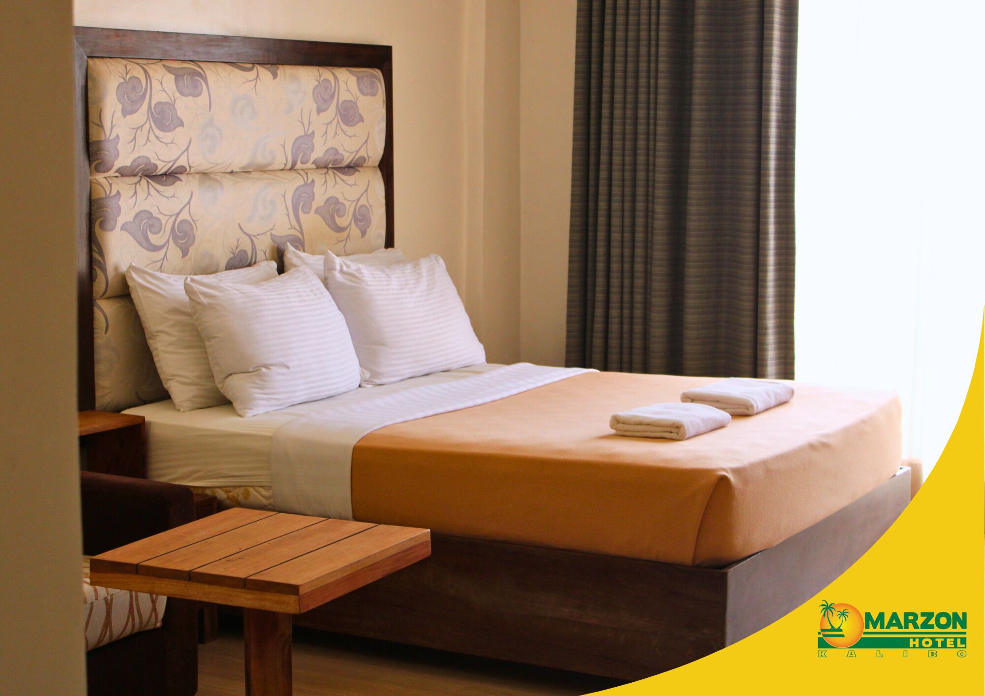 A hotel room with a bed, pillows, a wooden nightstand, and a table.