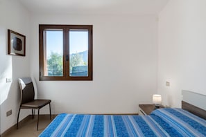 Une chambre à coucher comprenant un lit, une chaise, une table de chevet avec une lampe, une fenêtre donnant sur des arbres et un tableau encadré accroché au mur.