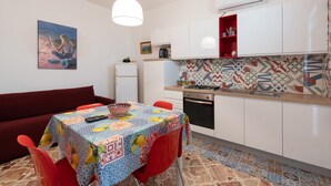 A compact kitchen with white cabinets, a red shelf, a patterned backsplash, and a dining area with a table and chairs. - Apartment 'Casa Rossa' with Sea View, Veranda & Wi-Fi (Castellammare del Golfio)