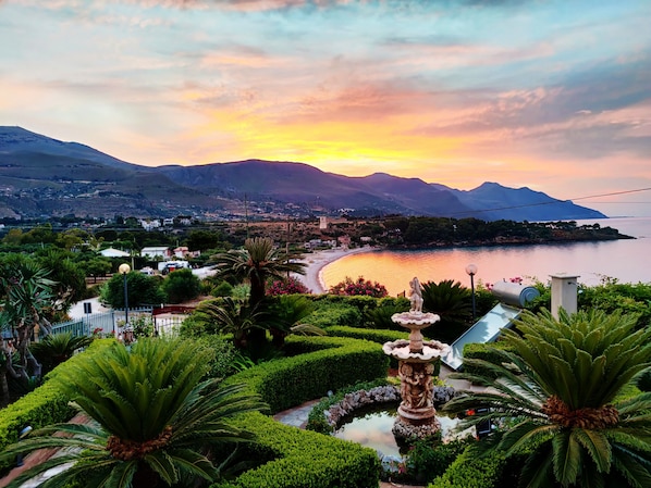 Un paysage côtier agrémenté d’une fontaine, de jardins soignés et offrant une vue sur la mer et les montagnes.