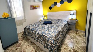 A bedroom with a patterned bedspread, a nightstand with a lamp, a wall-mounted air conditioner, and a window with blinds.