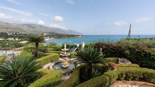 A landscaped garden with a fountain, palm trees, and a view of the sea.