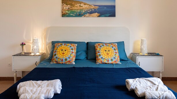 A bedroom with a bed, two decorative pillows, a nightstand with a lamp, and a painting of a coastal scene. - Apartment 'Mare & Relax' near the sea with terrace and Wi-Fi (Menfi)