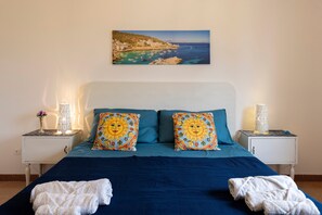 Une chambre à coucher avec un lit, deux coussins décoratifs, une table de chevet avec une lampe et un tableau représentant un paysage côtier.