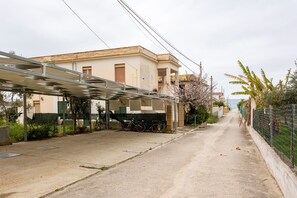Une rue étroite avec un espace de stationnement pour vélos abrité, un immeuble doté d’un balcon et une clôture ornée de verdure.