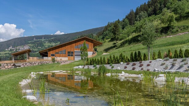 A modern house with a large wooden structure, a pond, and a hilly landscape in the background. - Holiday Apartment "Ringelblume Biohof Unterspisser" with Mountain View, Balcony & Wi-Fi (Lajen)