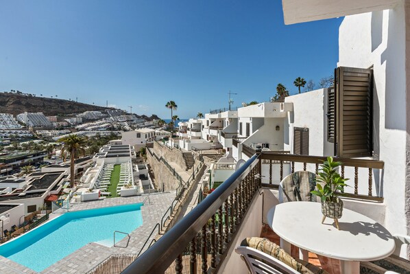 A rooftop pool with a view of a coastal town, a balcony with a table and chairs, and a small potted plant.