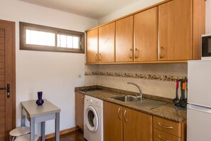 A compact kitchen with wooden cabinets, a washing machine, a sink, and a small table with a vase.