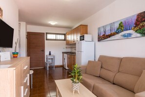 A living room with a sofa, a coffee table with a plant, a TV, a kitchen with cabinets, a washing machine, and a painting on the wall.