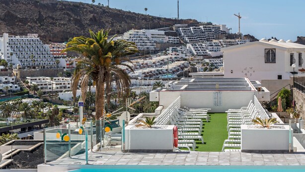 Una piscina en la azotea con vistas a un pueblo costero, incluyendo un hotel con balcones blancos y una palmera.