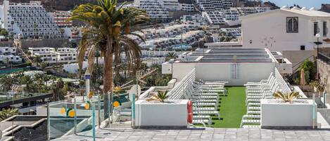 A rooftop pool with a view of a coastal town, including a hotel with white balconies and a palm tree.