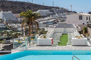A rooftop pool with a view of a coastal town, including a hotel with white balconies and a palm tree. - Apartamento Cumana 2 – Pool View, Mountain Views, Terrace, Shared Pool & Wi-Fi (Puerto Rico)