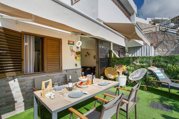 An outdoor dining area with a table set for two, surrounded by greenery and a view of a hillside.
