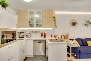 A modern kitchen with white cabinetry, a built-in oven, and a dining area with a blue sofa and yellow cushions.