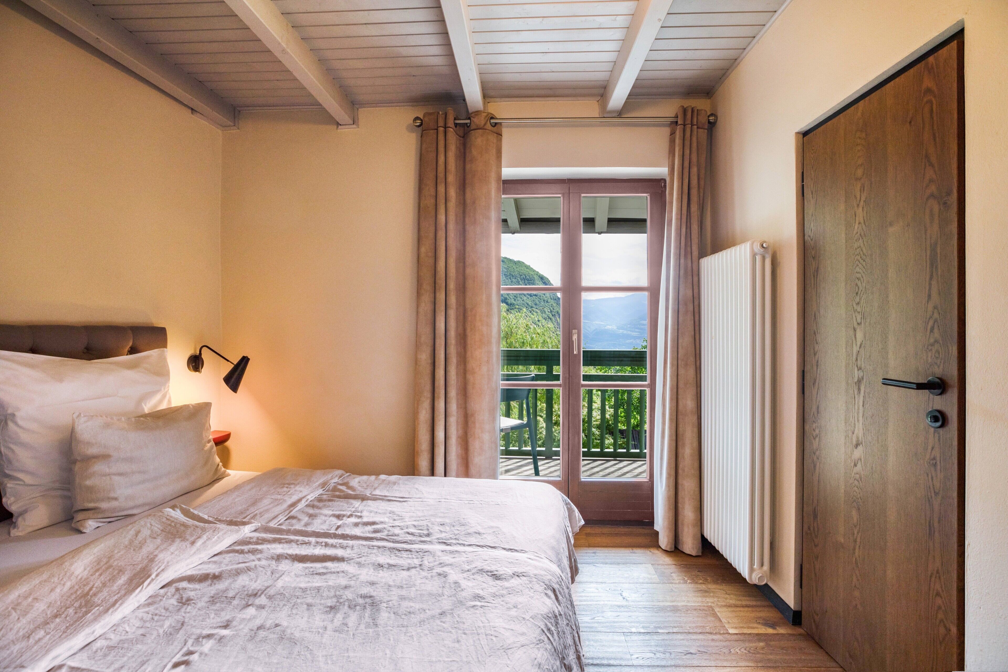 Ein Schlafzimmer mit Bett, Nachttisch, Wandlampe, Holztür und Blick auf einen Balkon mit einem Berg im Hintergrund.