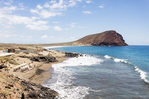 A beach with a rocky shoreline, a sandy area with sun loungers, and a prominent hill in the background.