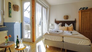 A bedroom with a bed, a wooden wardrobe, a small table with a bottle and glass, and a view of the outside.
