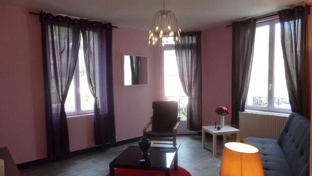 A modern living room with a sofa, armchair, and a coffee table. There is a lamp, a small table, and a window with curtains.