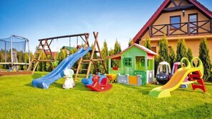 A playground with various play equipment including slides and swings, a small green house, and a large house with a red roof in the background. - Half of holiday home, terrace, playground (Rowy)