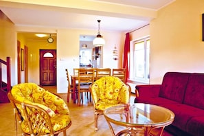 A living room with a red sofa, yellow floral armchairs, a glass coffee table, and a dining area with wooden chairs and a white table. - Half of holiday home, terrace, playground (Rowy)