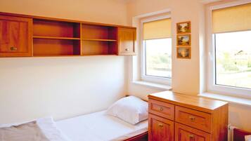 Une petite chambre avec un lit simple, une commode en bois, une fenêtre donnant sur un beau paysage et une étagère au-dessus du lit.