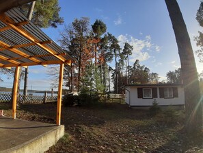 Un'area esterna coperta con una pergola in legno, un piccolo edificio bianco e un'area recintata con alberi e un lago sullo sfondo.