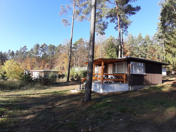 A cabin with a wooden deck and a covered porch, surrounded by tall trees and a clear sky.