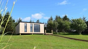 Eine moderne Holzbligge mit großen Fenstern, gelegen auf einem grasbewachsenen Hügel mit Bäumen im Hintergrund.