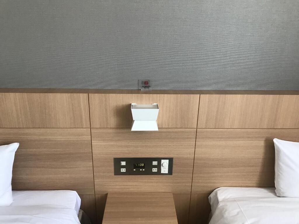 Une chambre d’hôtel avec une tête de lit en bois, une horloge numérique et une petite table placée entre deux lits.