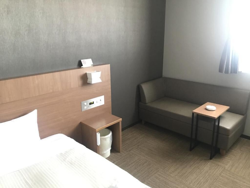 A hotel room with a bed, a small bedside table with a lamp, a chair, and a window with curtains.