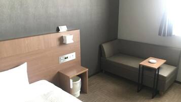 A hotel room with a bed, a small bedside table with a lamp, a chair, and a window with curtains.