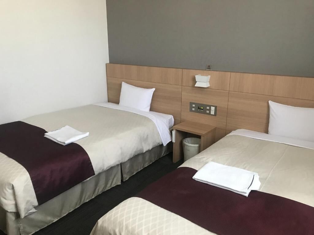 A hotel room with two beds, each with a white pillow and a dark red blanket. There is a wooden headboard with a control panel and a small trash bin.