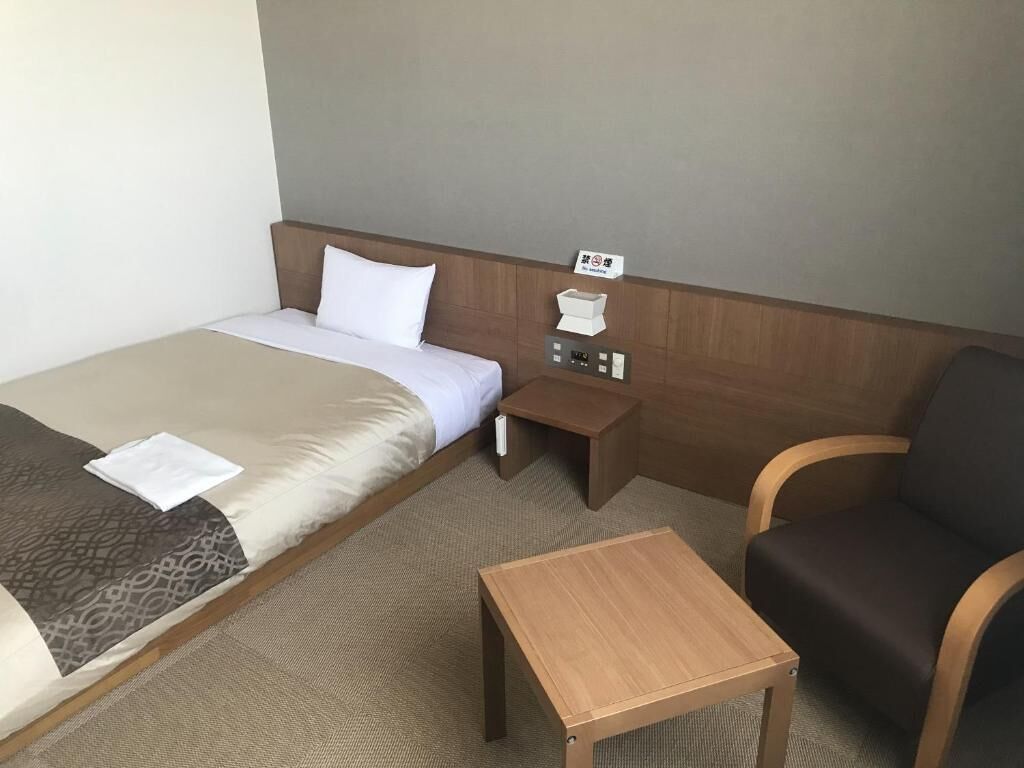 A hotel room with a single bed, a small wooden nightstand, a bedside table, a chair, and a wall-mounted control panel.