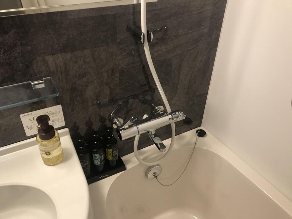 Un baño con bañera, ducha y lavamanos. Hay artículos de aseo en el lavamanos.