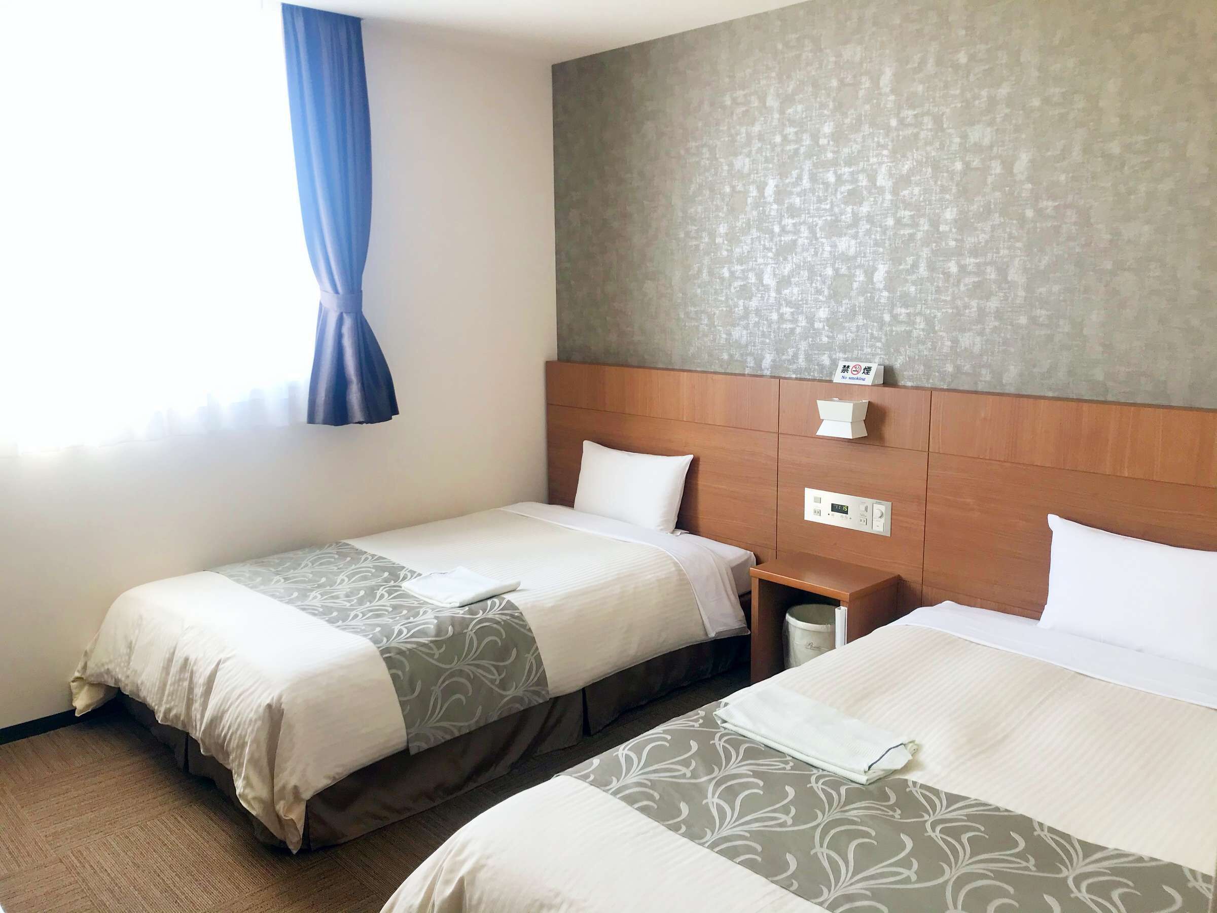 A hotel room with two beds, a wooden headboard, a bedside table with a lamp, and a wall-mounted thermostat.
