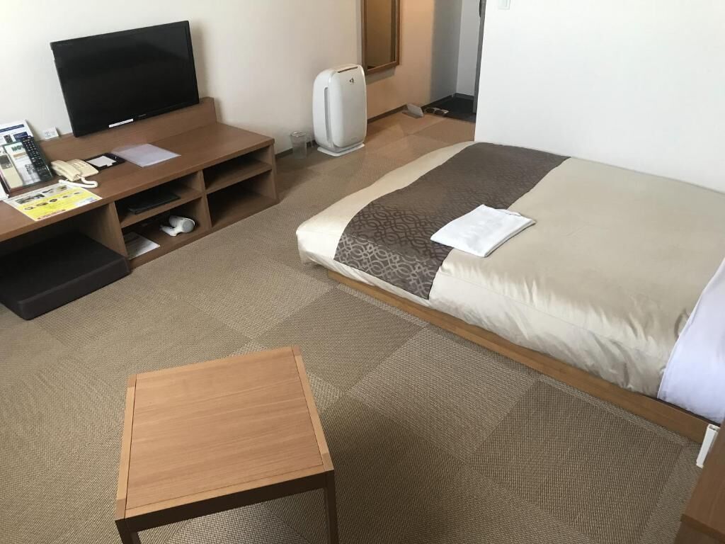 A hotel room with a bed, a wooden desk, a television, a heater, and a small table.