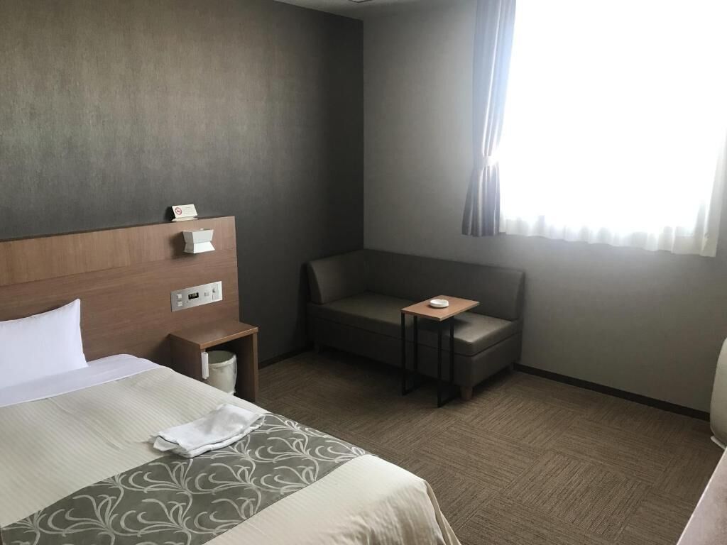 Ein Hotelzimmer mit Bett, einer kleinen Couch, einem Nachttisch und einem Fenster mit Vorhängen.