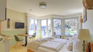 A hotel room with a bed, a desk, a chair, a TV, and large windows. - Apartment with balcony directly on the banks and with a view of the Moselle (Bullay)