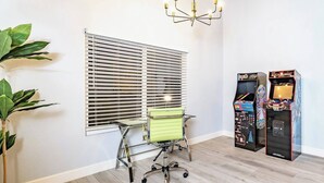 A room with a desk, two arcade games, a green chair, and a window with blinds.