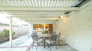 An outdoor patio with a round table and chairs, a pergola overhead, and a view into the house through large windows.