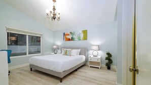 A bedroom with a large bed, a chandelier, a window, a bedside table with a lamp, and a painting on the wall.